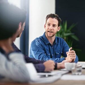 Homme en chemise bleue discutant lors d’une réunion professionnelle autour d’une table de bureau.